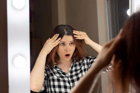 Horrified young woman looking in mirror staring open mouthed at the first grey hair on her scalp, a first sign of ageing.の写真素材