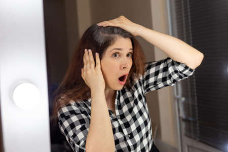 Horrified young woman looking in mirror staring open mouthed at the first grey hair on her scalp, a first sign of ageing.の写真素材