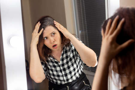 Horrified young woman looking in mirror staring open mouthed at the first grey hair, a first sign of...