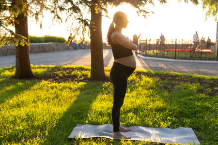 Side view of young woman expecting baby in black suit stands on rug barefoot and meditates on warm sunny summer day. Concept of sports and meditation activitiesの写真素材