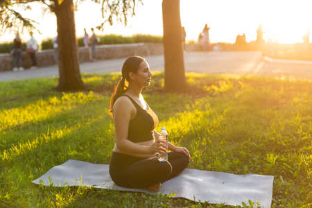 Cute young caucasian pregnant woman is meditating while sitting on a rug on the lawn on a sunny...