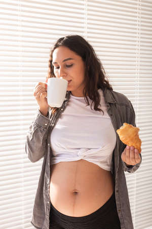 Healthy beautiful pregnant woman drinking tea and eating croissant during lunch. Concept of high-calorie nutrition while waiting for birth of babyの写真素材