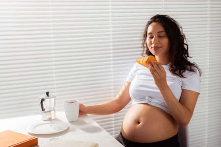 Joyful pregnant young beautiful woman eating croissant and drinking tea during morning breakfast. Concept of pleasant morning and expectation of meeting baby. Copyspaceの写真素材