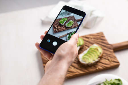 Hands take pictures on smartphone of two beautiful healthy sour cream and avocado sandwiches lying on board on the table. Social media and food conceptの写真素材