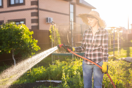 Caucasian woman gardener in work clothes watering the beds in her vegetable garden on sunny warm summer day. Concept of working in the garden and your farmの写真素材