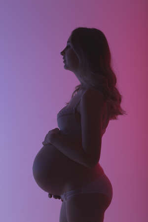 Beautiful young pregnant woman in lingerie is happily stroking her belly in studio in beautiful neon purple lighting. Concept of beauty and expectation of meeting with your beloved babyの写真素材