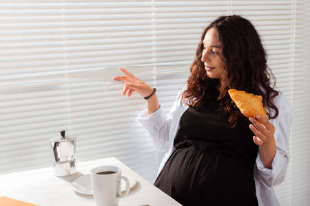 Happy pregnant young beautiful woman eating croissant and looking througt blinds during morning breakfast. Concept of pleasant morning and positive attitude during pregnancyの写真素材