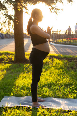 Side view of young woman expecting baby in black suit stands on rug barefoot and meditates on warm sunny summer day. Concept of sports and meditation activitiesの写真素材