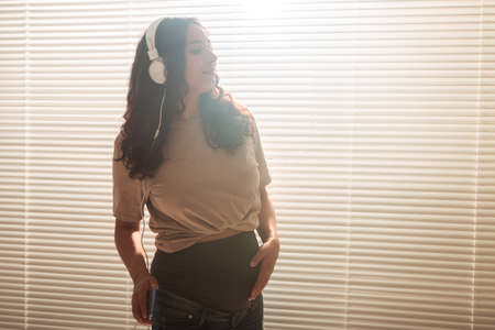Curly-haired brunette pacified pregnant woman listens to pleasant classical music using smartphone and headphones. Concept of a soothing mood before meeting baby. Copyspaceの写真素材