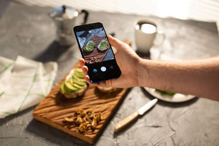 Hands take pictures on smartphone of two beautiful healthy sour cream and avocado sandwiches lying on board on the table. Social media and food conceptの写真素材