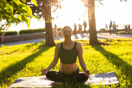 Cute young caucasian pregnant woman is meditating while sitting on a rug on the lawn on a sunny summer evening. Concept of pacification and energy boostの写真素材