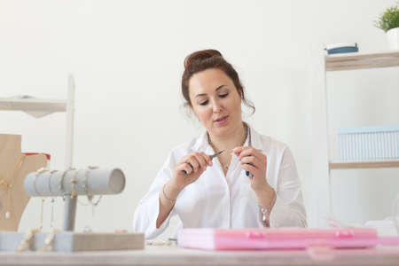 Charming young enthusiastic caucasian woman making beautiful unique costume jewelry while sitting at her desk. Concept of hobby and pleasant workの写真素材