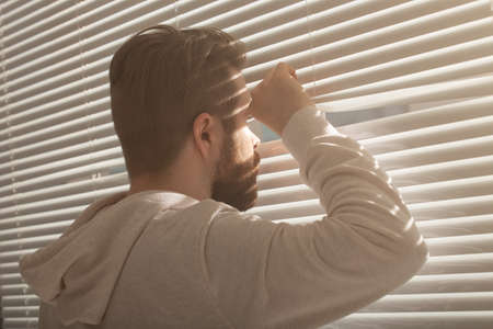 Rear view of young man with beard peeks through hole in the window blinds and looks out into the street. Surveillance and curiosity conceptの写真素材