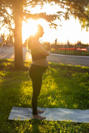 Side view of young woman expecting baby in black suit stands on rug barefoot and meditates on warm sunny summer day. Concept of sports and meditation activitiesの写真素材