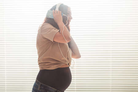 Curly-haired brunette pacified pregnant woman listens to pleasant classical music using smartphone and headphones. Concept of a soothing mood before meeting baby. Copyspaceの写真素材