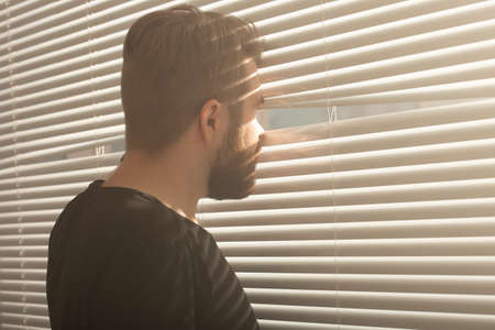 Rear view of young man with beard peeks through hole in the window blinds and looks out into the street. Surveillance and curiosity conceptの写真素材
