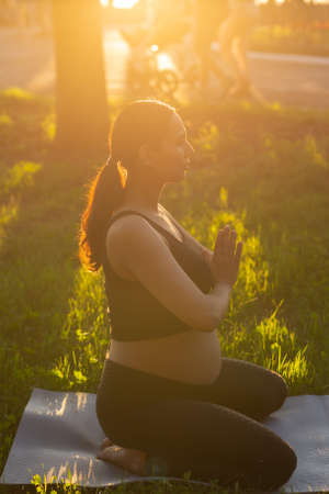 Cute young caucasian pregnant woman is meditating while sitting on a rug on the lawn on a sunny summer evening. Concept of pacification and energy boostの写真素材