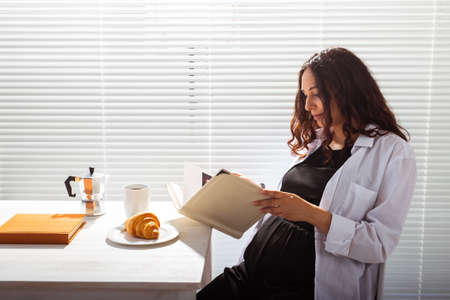 Side view of happy pregnant woman reading book while having morning breakfast with coffee and...