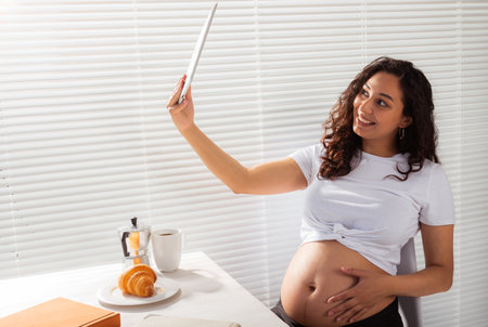 Happy pregnant young beautiful woman talking to mom using video call during morning breakfast. Concept of communication and positive attitude during pregnancyの写真素材