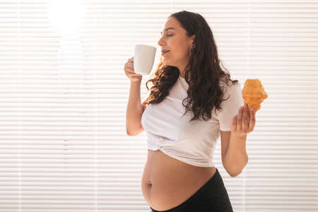 Beautiful pregnant woman holding croissant and cup of coffee in her hands during morning breakfast. Concept of good health and positive attitude while expecting baby. Copy space.の写真素材