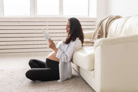 Pregnant woman holding tablet sitting on a carpet near a couch in the living room at homeの写真素材