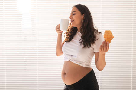 Happy pregnant woman with croissant buns at home. Pregnancy, eating and people conceptの写真素材