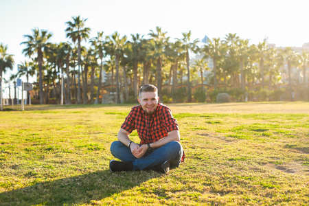 Handsome young smiling man sitting on the grass at park. Summer in the city concept.