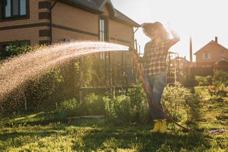 Happy woman gardener in work clothes watering the beds in her vegetable garden on sunny warm summer day. Concept of working in vegetable garden on your farmの写真素材