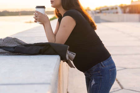 Young pregnant woman drinking takeaway coffee or tea on embankment. Maternity, leisure and health concept.の写真素材
