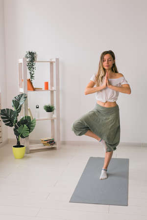 Woman practise yoga performs. Tree pose standing on mat inside of cozy room with plants and...
