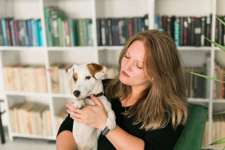 Happy female owner of jack russell terrier dog, feels responsibility of caring about pet, standing against bookshelves background. People and relationship with animalsの写真素材