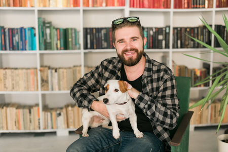 Happy male owner of jack russell terrier dog, feels responsibility of caring about pet, standing against bookshelves background. People and relationship with animalsの写真素材
