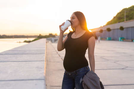 Young pregnant woman drinking takeaway coffee or tea on embankment. Maternity, leisure and health concept. Advertising area workspaceの写真素材