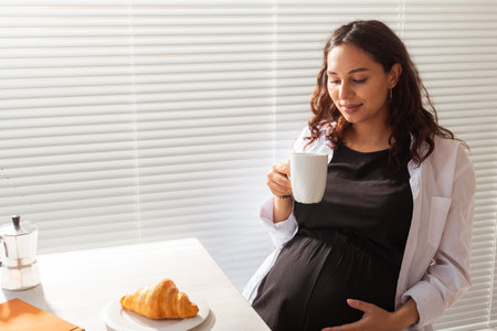 Happy pregnant young beautiful woman drinks tea during morning breakfast. Concept of pleasant morning and positive attitude during pregnancyの写真素材