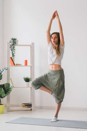 Woman practise yoga performs. Tree pose standing on mat inside of cozy room with plants and greenery. Healthy lifestyle concept. Copy space.の写真素材