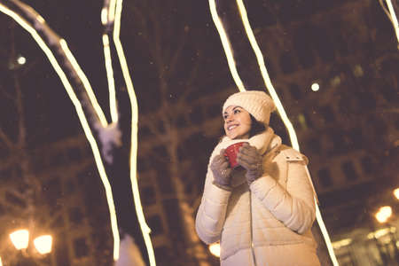 Girl standing over winter christmas city background snow snow drifts, stands warm jacket hat, holds cup tea with hot tea drinks.の写真素材