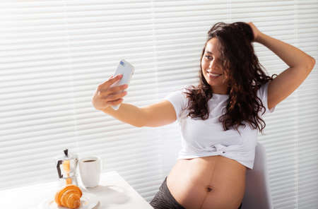 Pregnant woman takes selfie on her smartphone while breakfast. Maternity leave and technologies concept.の写真素材