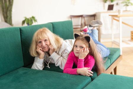 Happy granddaughter and grandmother lying on sofa in funny pose. Grandma having fun with grandchild at home. Senior people, family and generation concept.の写真素材