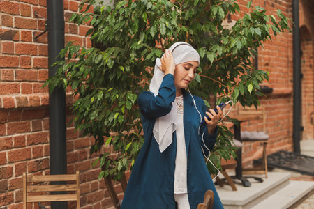 Portrait of young modern arabian woman holding mobile phone and listening the music to headphone.の写真素材