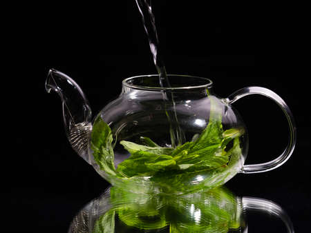 Pouring hot water into a glass teapot on a black background, green mint tea. Herbal tea and healthy drink concept.の写真素材