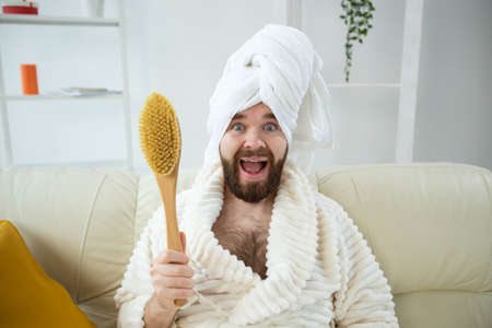 Portrait of funny guy wear turban towel holding in hands massage brush while sits on sofa. Male skin care and spa concept.の写真素材