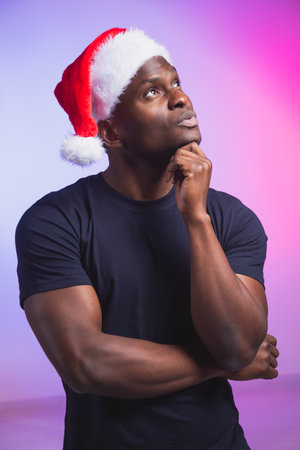 Portrait of pensive african american man in santa hat and casual t-shirt on colourful background, christmas time. Winter holidays and emotional people concept.の写真素材