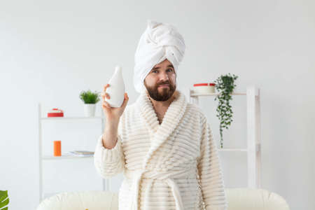 Beauty portrait of handsome beautiful guy in towel and bathrobe holding tube of body cream lotion in...