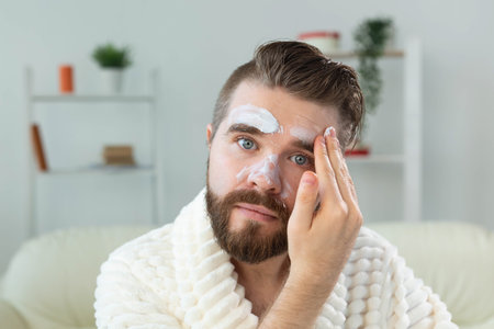 Bearded guy applying anti-wrinkle cream on his face. Skin care and spa for man concept.の写真素材