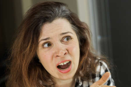 Horrified young woman looking in the bathroom mirror staring open mouthed at the first grey hair on...