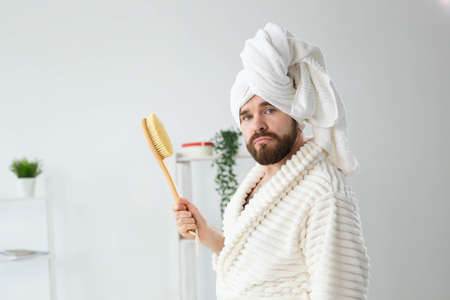 Portrait of funny guy wear turban towel holding in hands massage brush while sits on sofa. Male skin care and spa concept.の写真素材