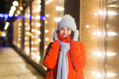 Girl having fun on christmas decoration lights street. Young happy smiling woman wearing stylish knitted scarf and jacket outdoors. Model laughing. Winter wonderland city scene, New Year party.の写真素材