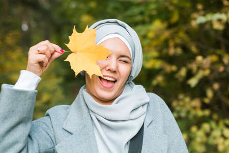 Autumn muslim woman holding maple leaf outdoors in sunny day. Beautiful arab wearing hijab girl in...