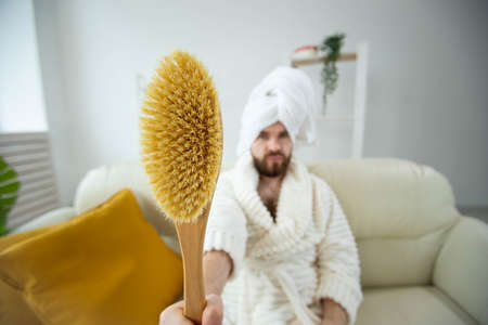 Portrait of funny guy wear turban towel holding in hands massage brush while sits on sofa. Male skin care and spa concept.の写真素材