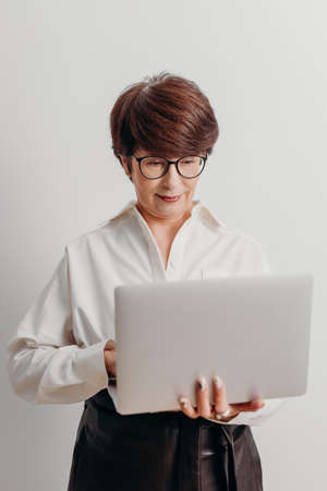 Positive middle aged business woman in white blouse touches pad of laptop standing near brick wall in office.の写真素材